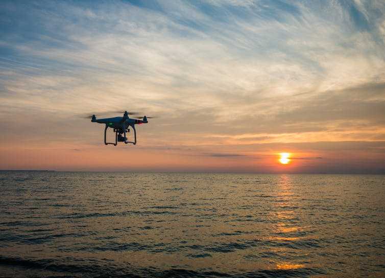 A drone is silhouetted against a sunset, with the sea below
